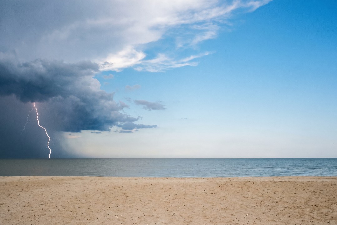 A lightning bolt strikes the ocean on the left of the frame beneath an isolated dark storm cell, while the sky on the right and overhead is bright and clear blue.