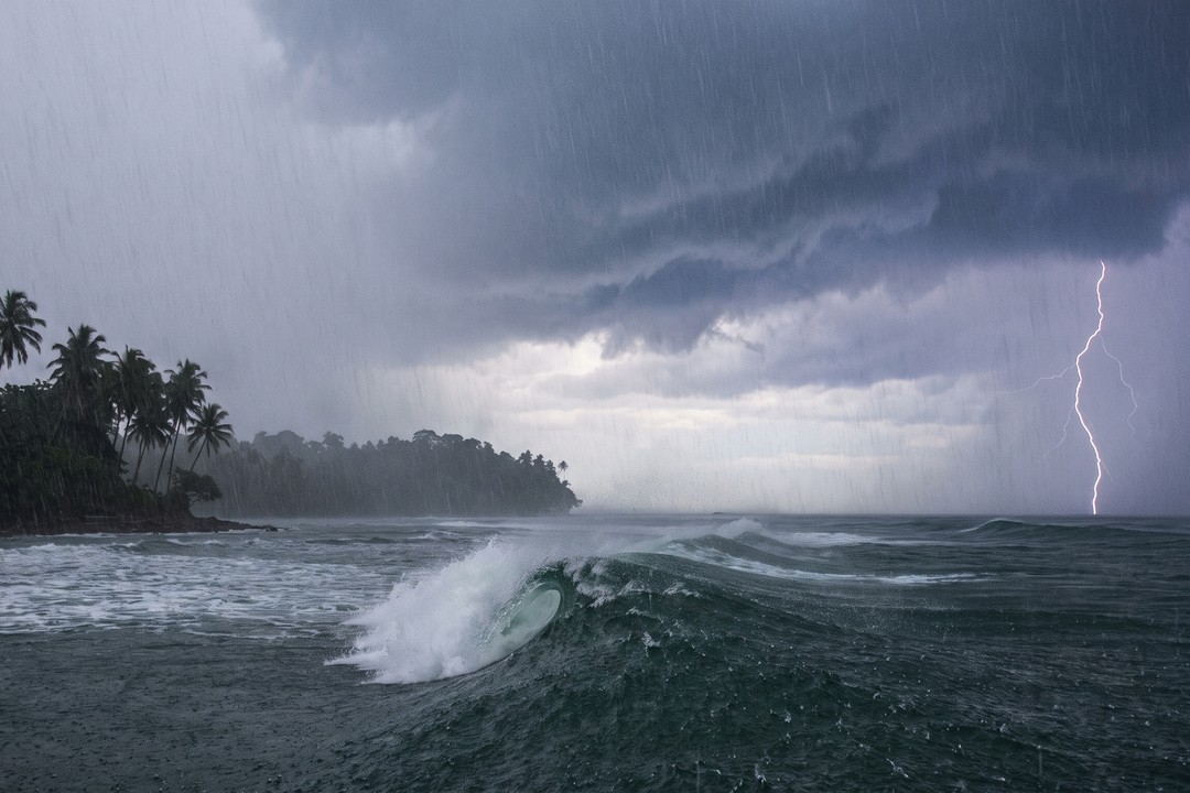 A branching lightning bolt strikes the open ocean beneath a dark storm sky, its channel reflected on the wet sand of an empty beach in the foreground.