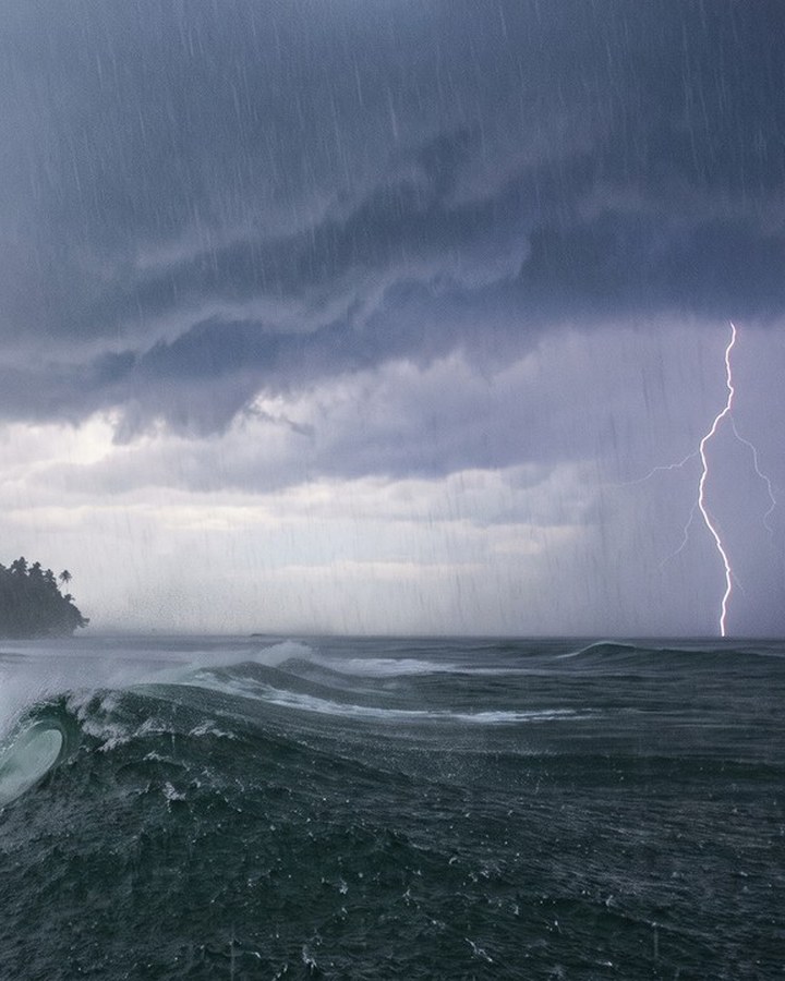 A surfer sitting alone on a board in gray water beneath a low, dark storm front, waiting between sets.