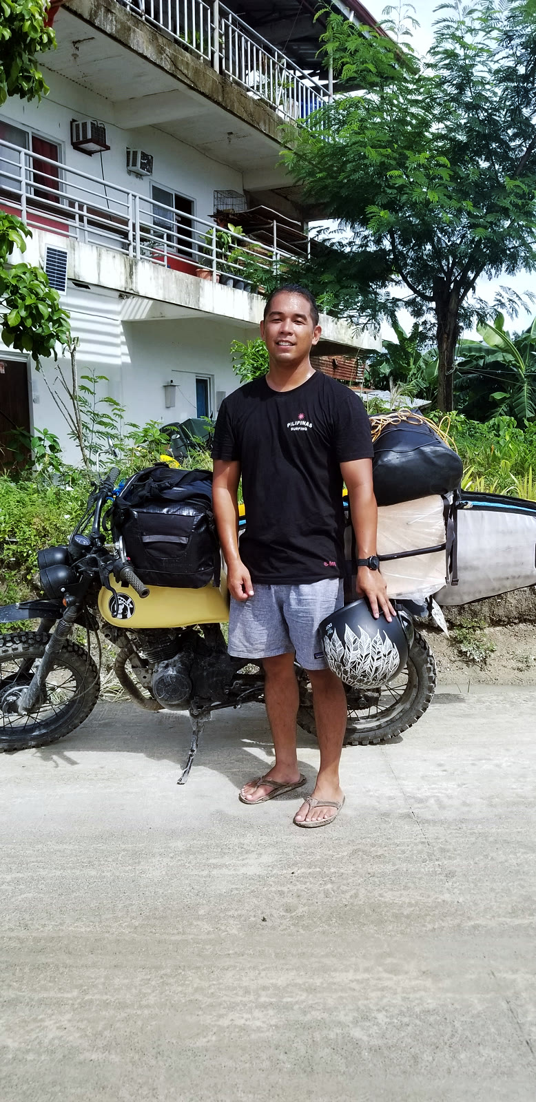 Dr. Divinagracia standing beside a yellow dual-sport motorcycle loaded with a surfboard, dry bags, and a helmet.