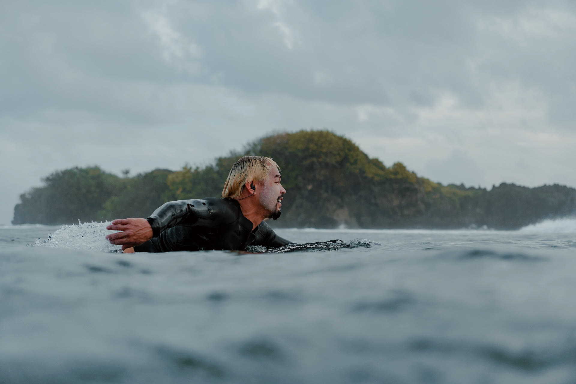 Side-profile water-level shot of Dr. Divinagracia paddling out, ear plugs visible, forested rocky island in the background. The surfer the medicine comes from.