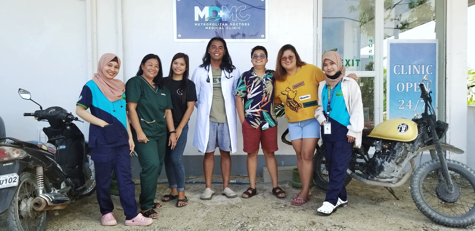 Dr. Divinagracia in a white coat with a stethoscope, standing with six clinic staff outside Metropolitan Doctors Medical Clinic.