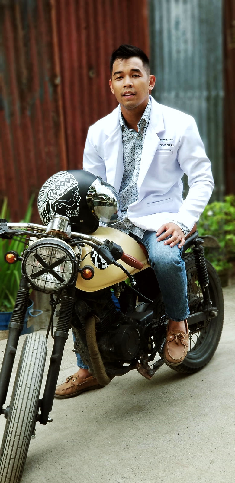 Dr. Divinagracia in a white coat seated on a yellow motorcycle, helmet resting on the handlebars, in front of a rust-textured metal wall.