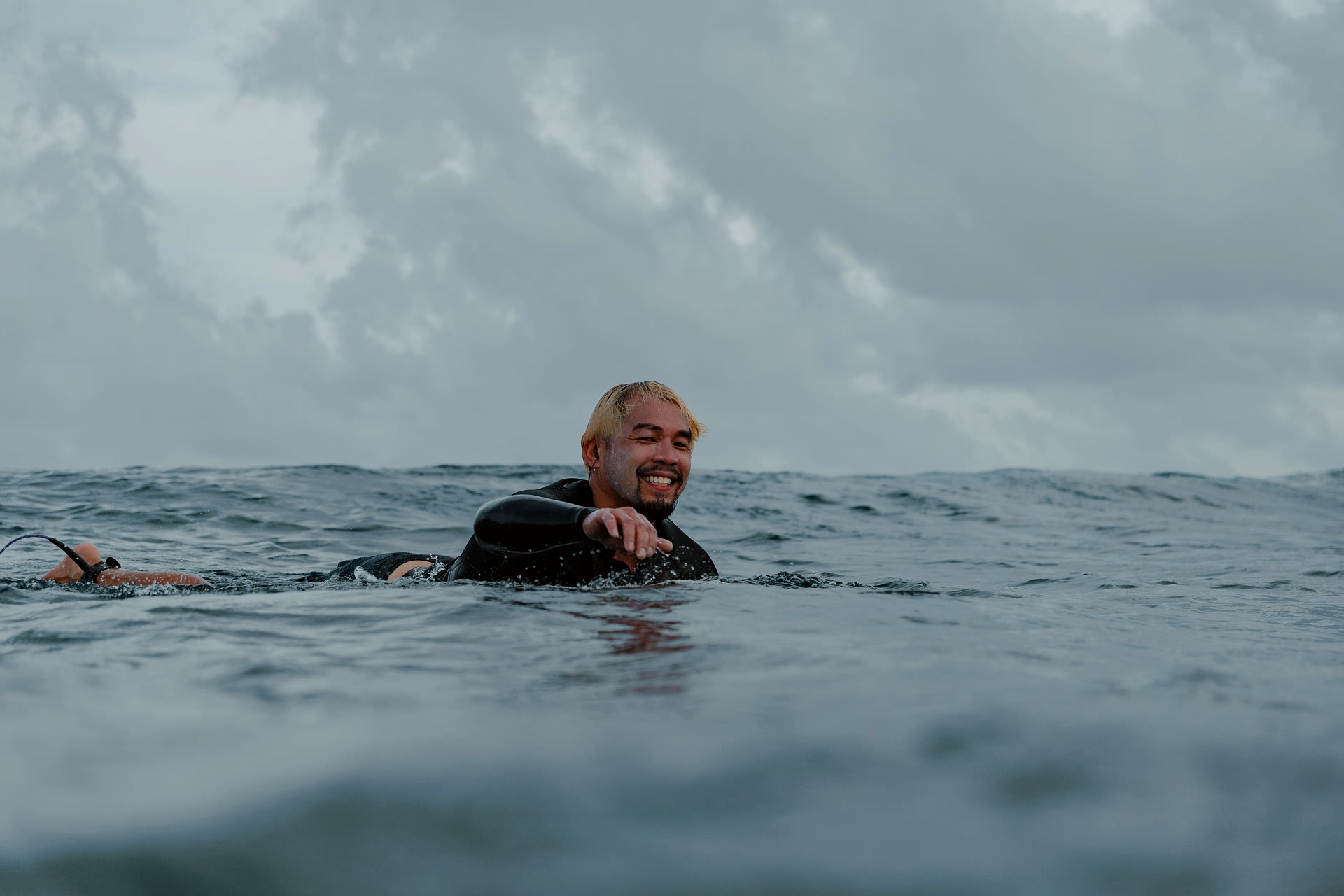 Dr. Divinagracia smiling mid-paddle-out on a black surfboard, water-level shot, moody clouded sky above the open ocean.