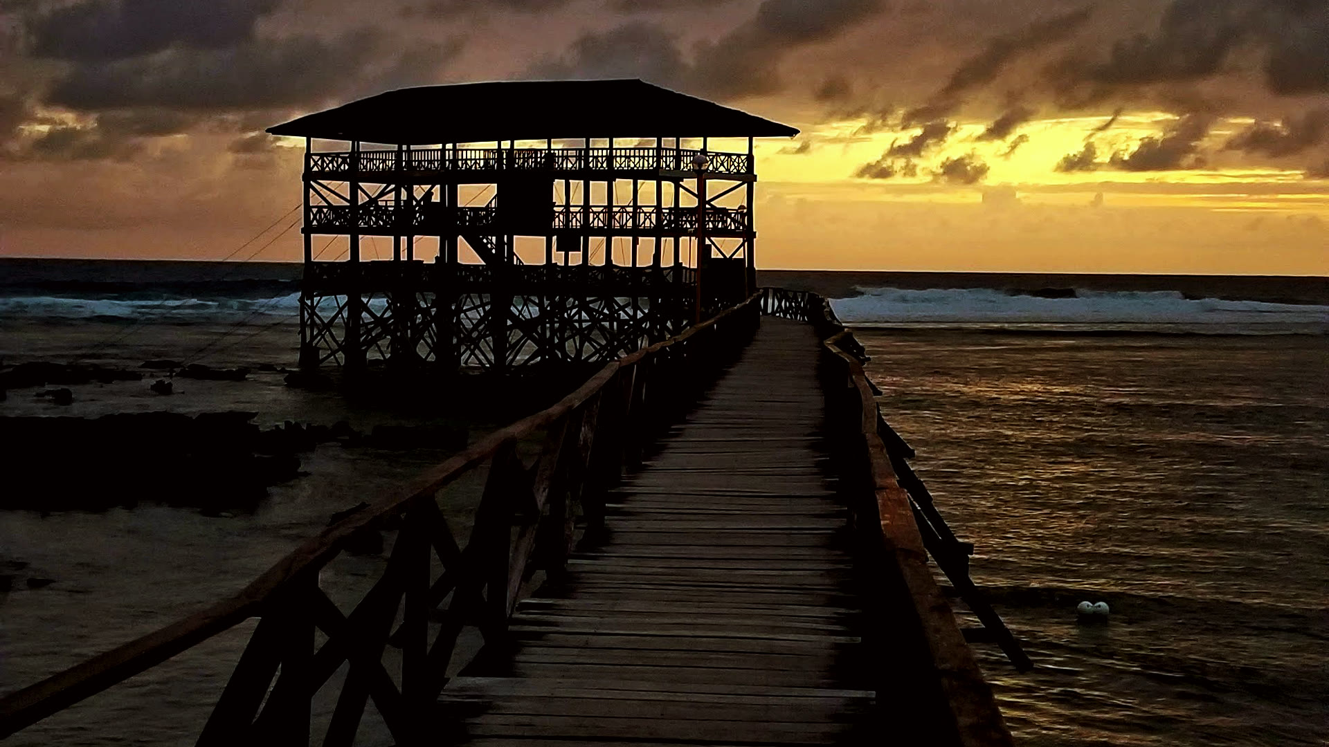 The Cloud 9 boardwalk in Siargao leading out to the iconic tower at sunset, with dramatic clouds reflected on the wet wood.