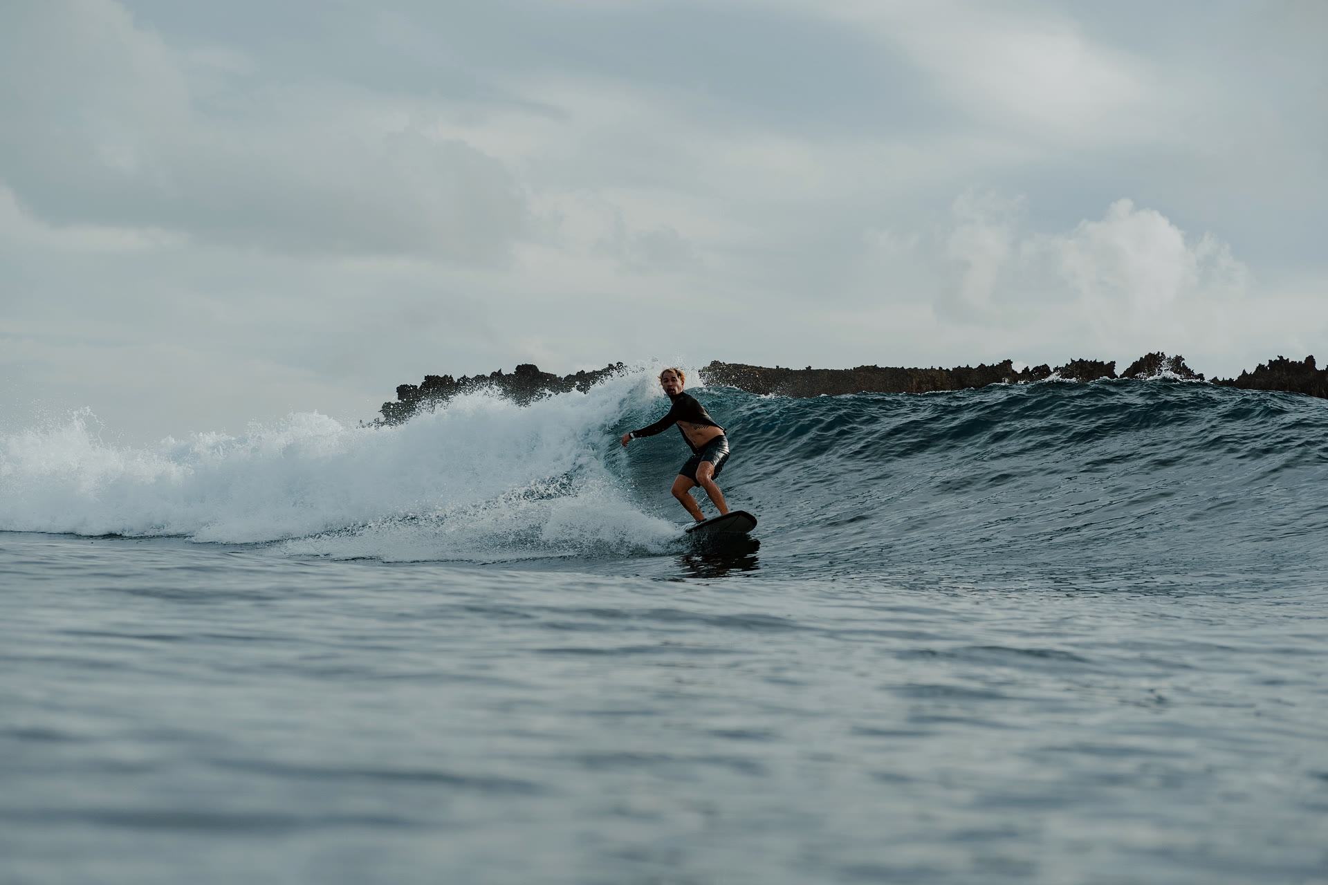 A surfer carves a wave with a rocky coastline visible in the background, water-level documentary photograph.